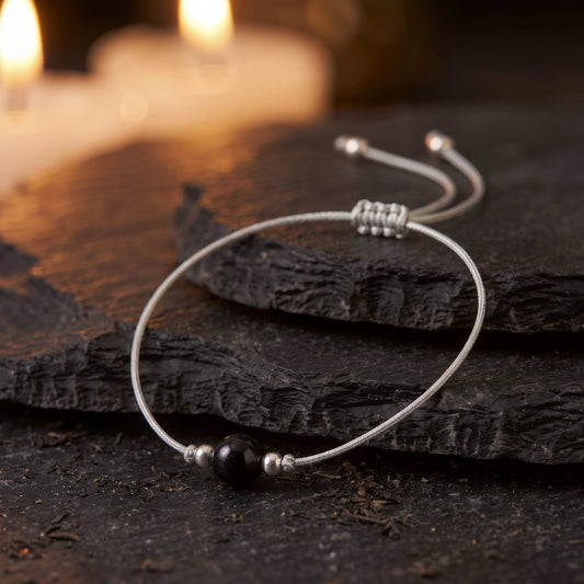 Pulsera Amuleto Turmalina Negra en hilo gris ajustable sobre una piedra oscura, con velas al fondo